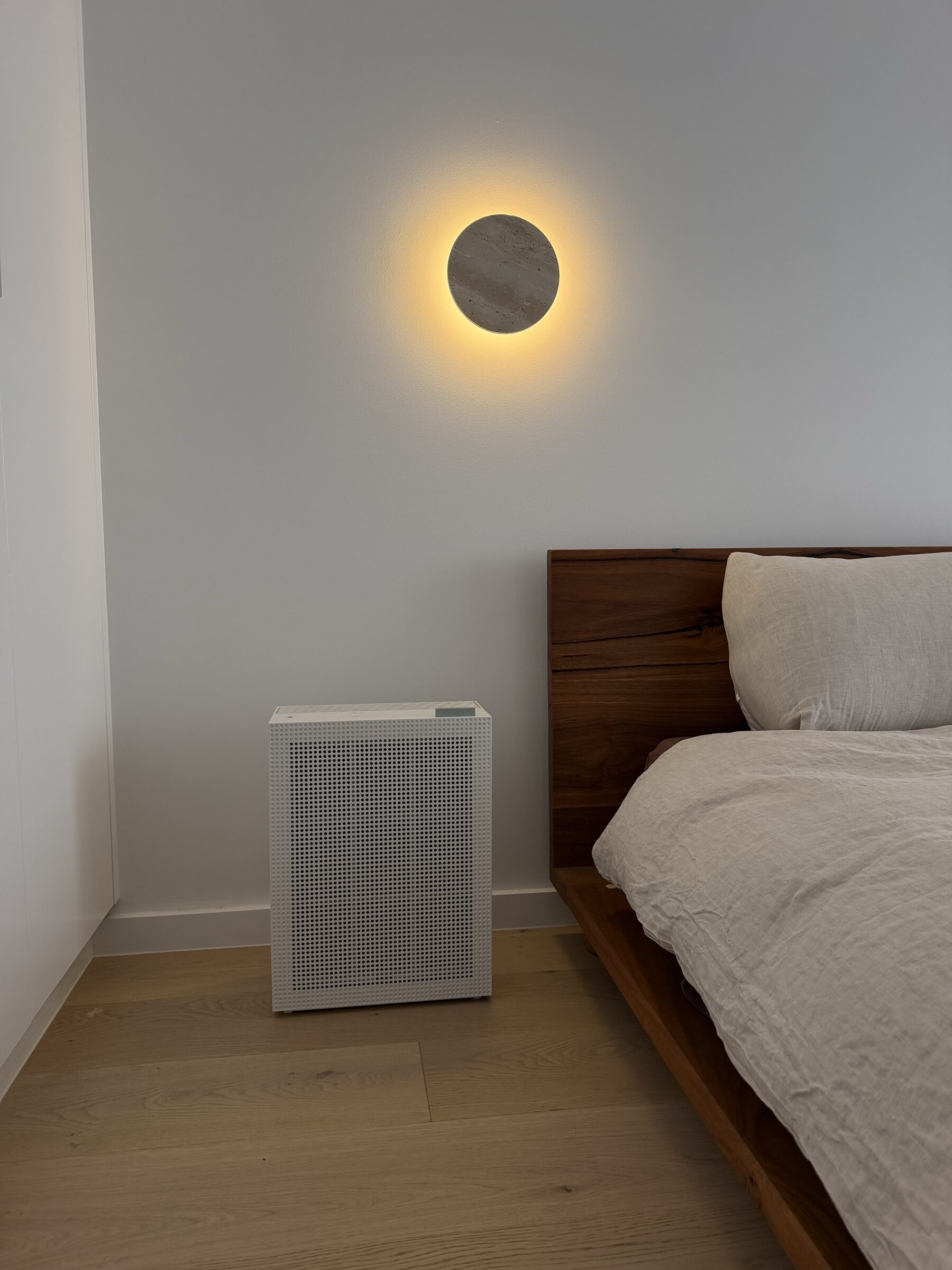 Coway Airmega 150 at the side of a timber bed under a circular travertine wall sconce